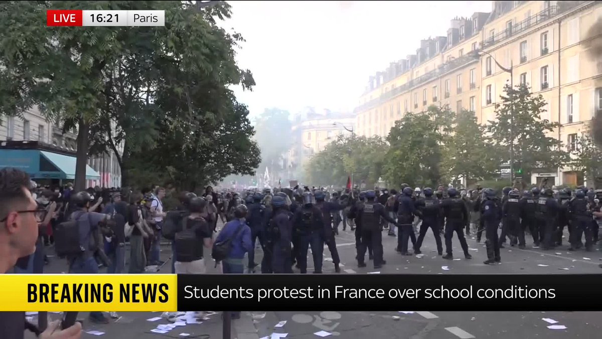A live breaking news broadcast from Paris showing a tense standoff between a large crowd of protesters and police in riot gear on a city street.