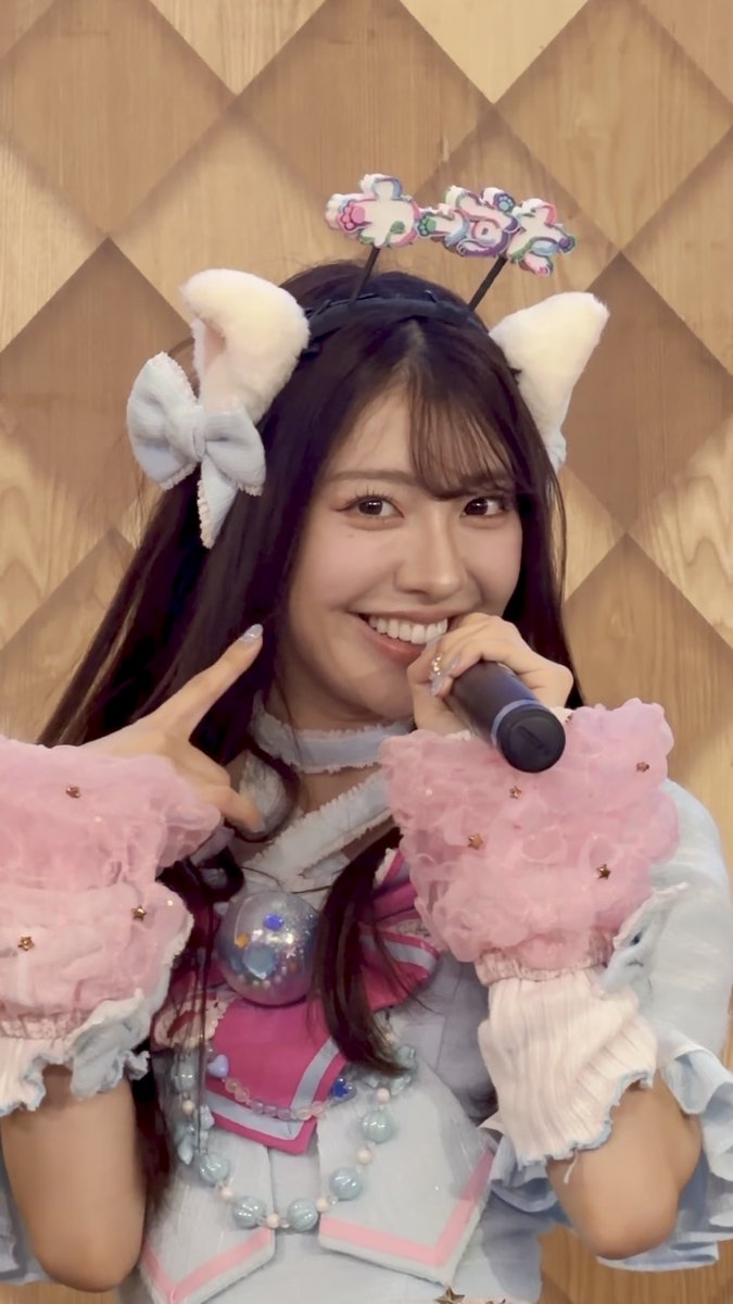 A smiling young woman with long dark hair wears cat ears and a pastel idol-style outfit with a bear-shaped headband, holding a microphone and making a peace sign.