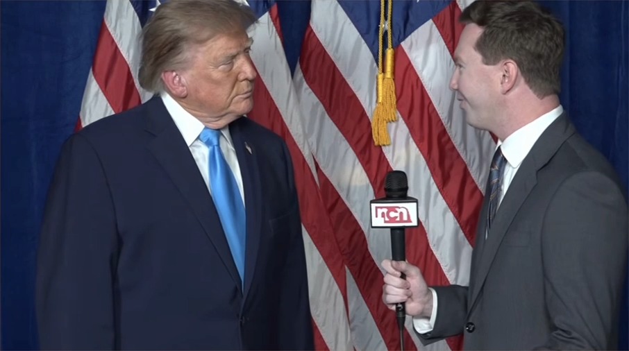 A man in a blue suit and blue tie stands face-to-face with a reporter holding an 'NCRH' microphone in front of an American flag, appearing to be interviewed.
