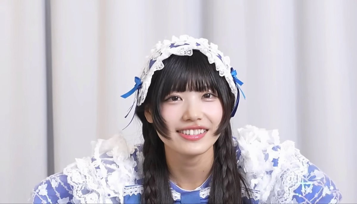 A smiling young woman with dark hair wearing a white lace headpiece with blue ribbons and an ornate blue and white lace outfit.