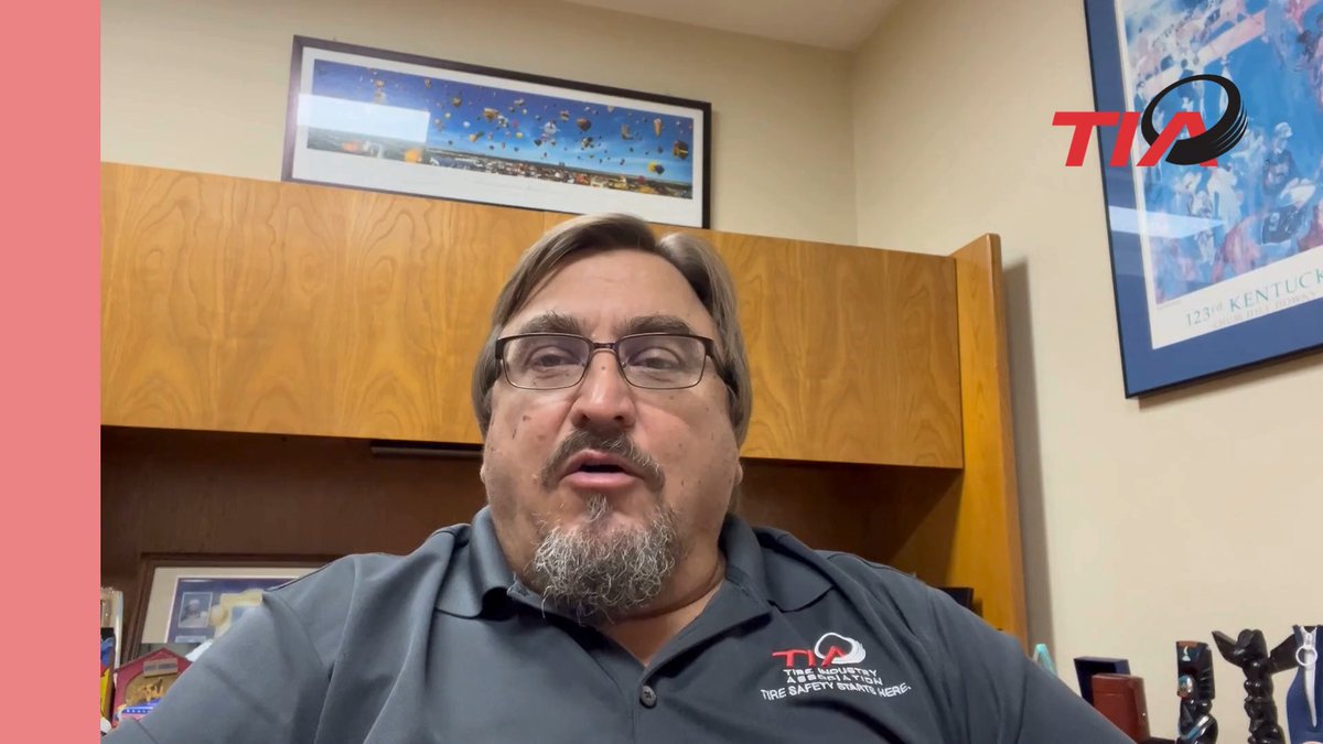 A man with glasses, a mustache, and goatee speaks to the camera, wearing a gray polo shirt with the logo 'TIA - Tire Industry Association - Tire Safety Starts Here' visible on the chest. Behind him is a wooden cabinet, framed pictures including a hot air balloon print and a '123rd KENTUCKY' poster, plus a 'TIA' watermark in the corner.