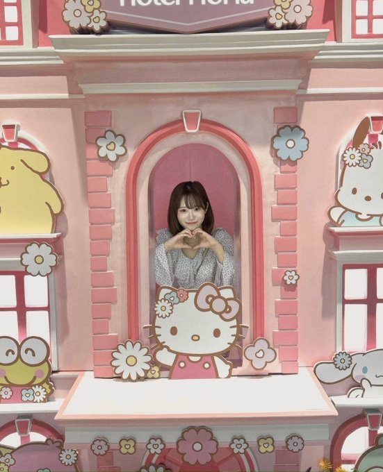 A young woman with long dark hair stands inside a pink Hello Kitty themed photo spot, making a heart shape with her hands, surrounded by cute Sanrio character cutouts and floral decorations.