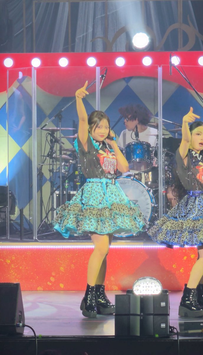 A young woman performs on stage in a blue and black outfit, pointing upward while singing, with another performer and a drummer visible behind her.