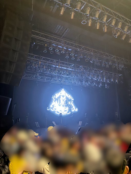 A concert stage is illuminated with blue lighting, featuring a bright diamond-shaped logo at the back, with the silhouettes of a crowd visible in the foreground.