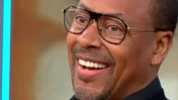 A close-up reaction shot of a man wearing glasses with an exaggerated, wide-eyed smile.
