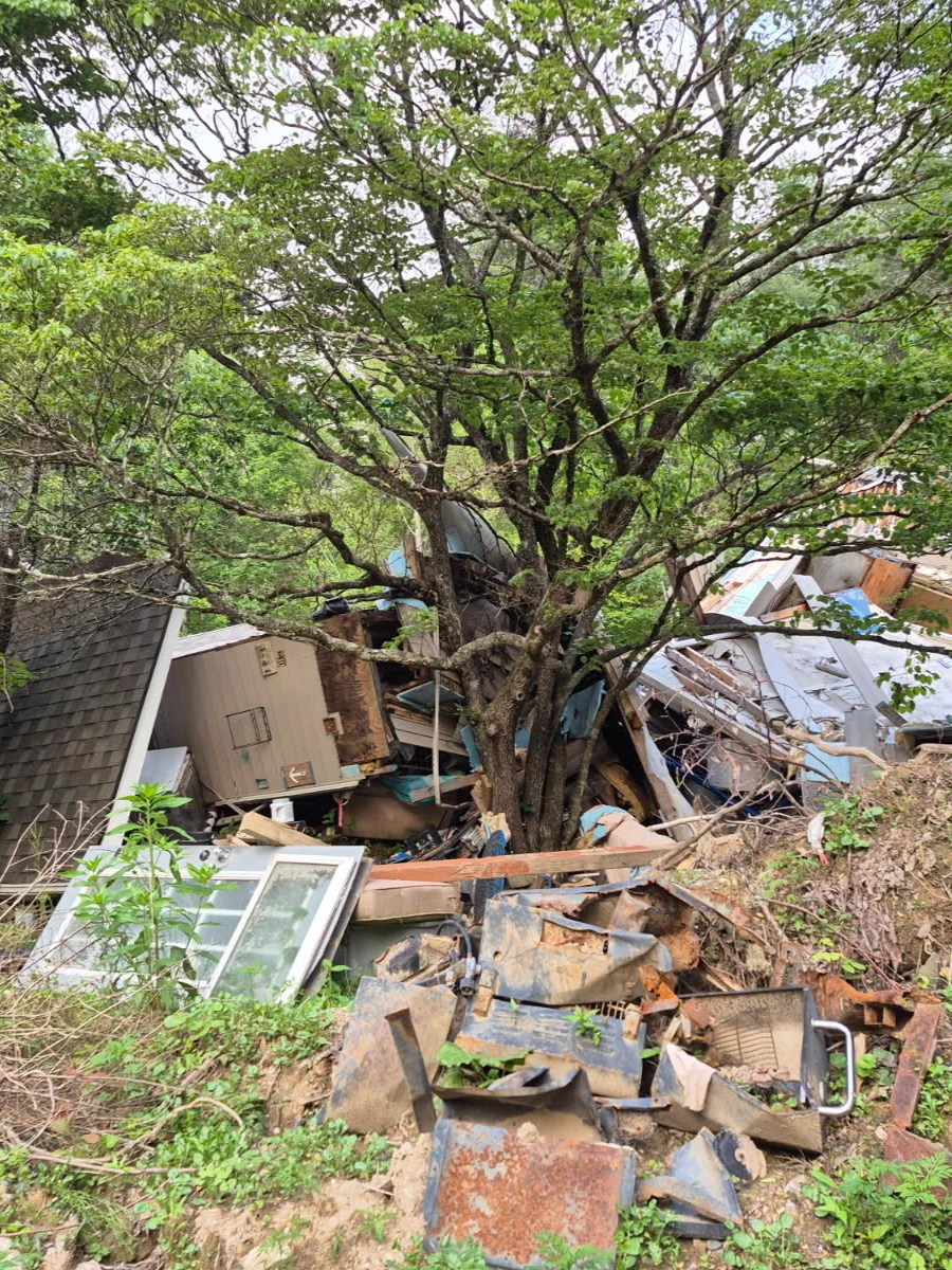 A collapsed house surrounded by trees, with various household debris and appliances scattered amidst green foliage.