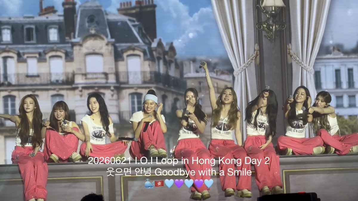 K-pop group performing at a Hong Kong concert on June 21, 2026, with members in matching outfits waving light sticks on stage with a Paris backdrop.