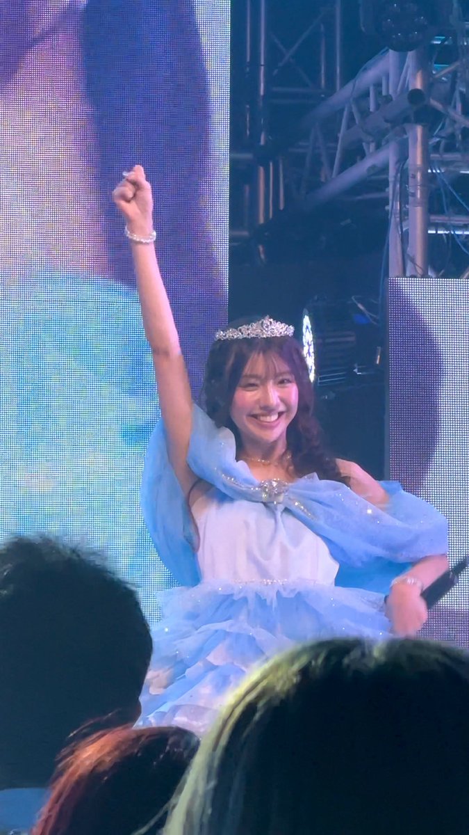 A young woman wearing a sparkly blue princess dress and tiara smiles and raises her fist on stage at a concert.