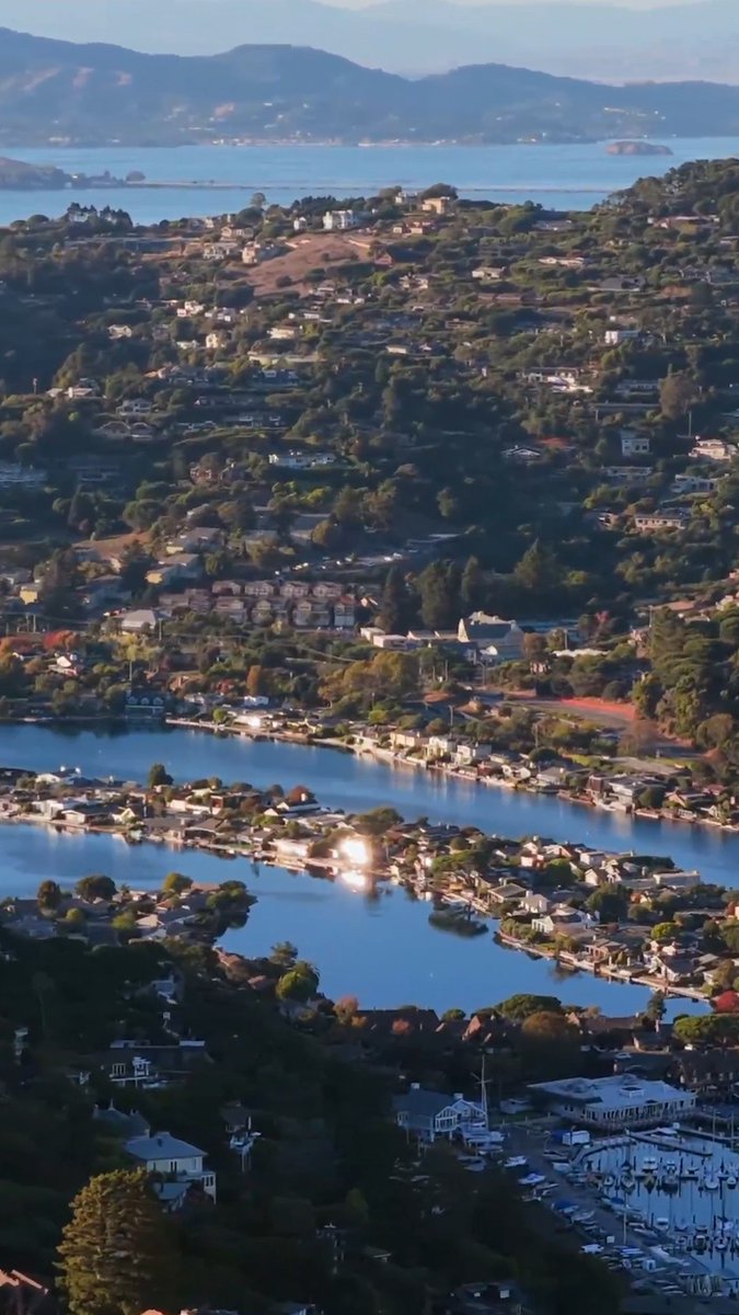 An aerial photograph of a coastal town with houses, a marina, and a lagoon in the foreground, with the San Francisco Bay visible in the distance.
