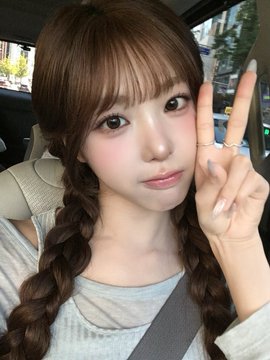 A young woman with braided brown hair and bangs takes a selfie in a car, flashing a peace sign while wearing a gray top and a seatbelt.