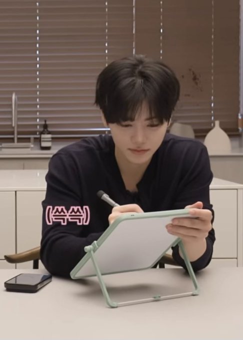 A young man in a dark shirt draws on a whiteboard with a stylus, with the pink text '(ㅅㅅ)' overlaid on his arm.