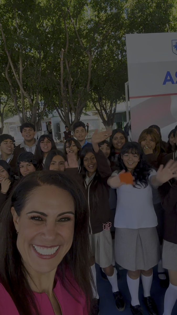 A selfie-style photo featuring a smiling woman in a pink jacket in the foreground, with a large group of smiling schoolgirls in brown and white uniforms gathered behind her, possibly at a school event.