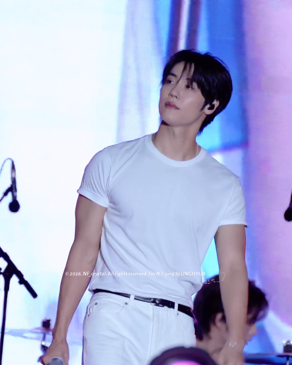 A performer with dark hair, wearing a fitted white t-shirt and white pants with a black belt, stands on stage with a contemplative expression, looking off to the side. A microphone stand and another band member are partially visible, while a watermark reads "© 2026. NF_crystal. All rights reserved. For N.Flying SEUNGHYUB," connecting the image to the post's mention of a live performance and comeback teaser.