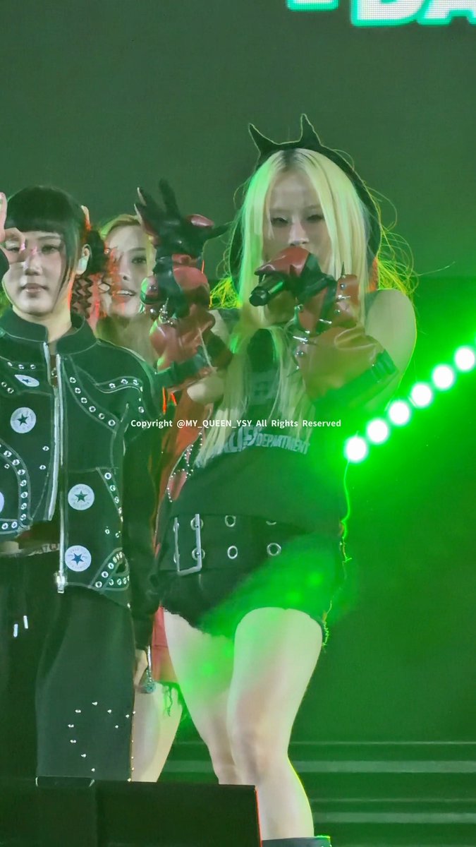 A performer with long blonde hair sings into a microphone on a stage illuminated by green lighting, wearing a black top and studded leather gloves; a watermark 'Copyright @MY_QUEEN_YSY All Rights Reserved' is visible.