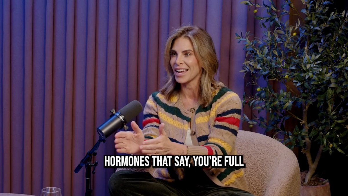 Jillian Michaels sits in a studio setting, gesturing animatedly while speaking into a microphone during a podcast episode about GLP-1 medications. The on-screen text "HORMONES THAT SAY, YOU'RE FULL" appears at the bottom, illustrating her explanation of how these drugs mimic satiety hormones to suppress appetite.