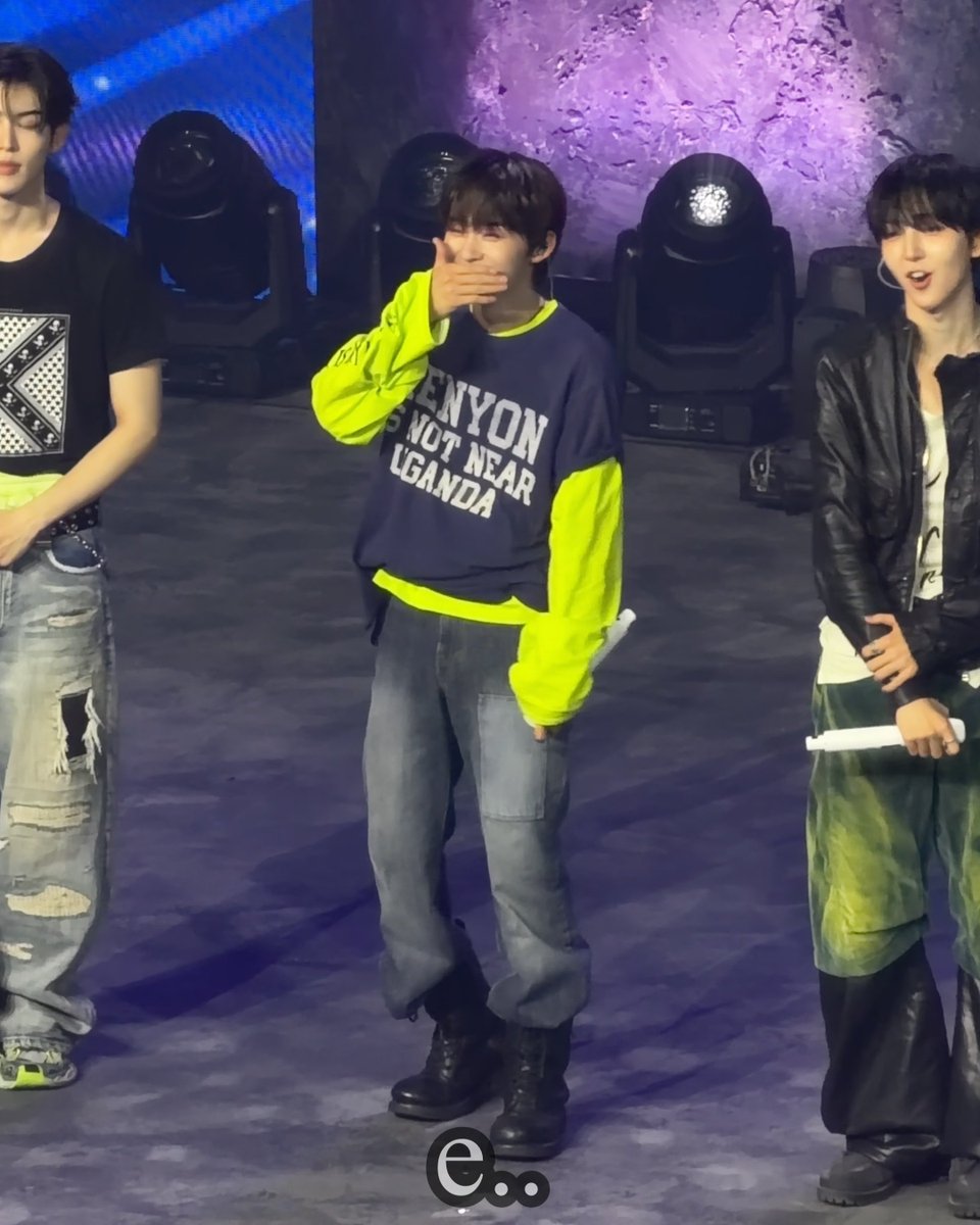 Three young men on a stage; the middle one wears a navy and neon green sweatshirt reading 'ENYON IS NOT NEAR LGANDA' and laughs while covering his mouth.