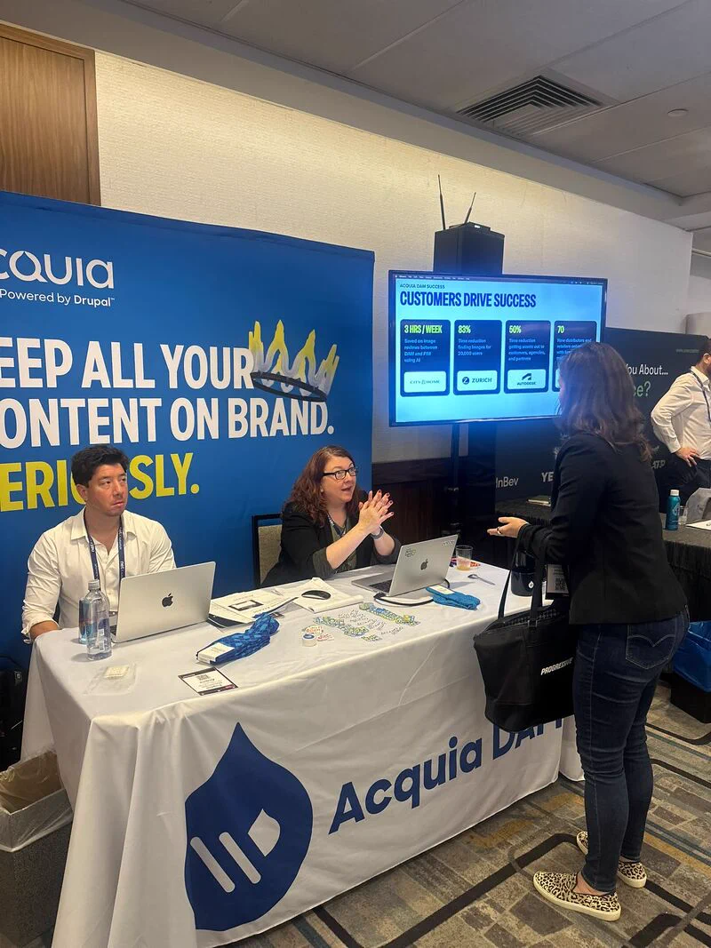 Two representatives at an Acquia booth engaging with a visitor during a conference.