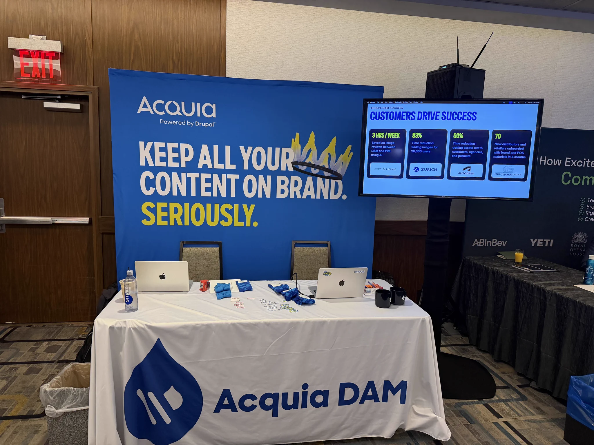 Acquia DAM booth with laptops and promotional items, featuring a banner emphasizing brand content control.