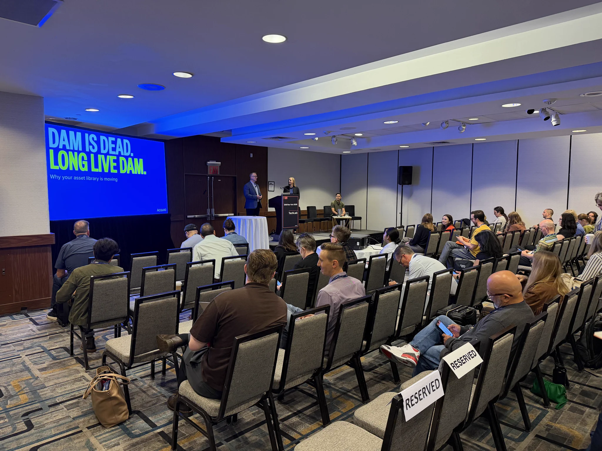 Audience seated in a conference room listening to a speaker presenting a slide titled 'DAM IS DEAD, LONG LIVE DAM.'