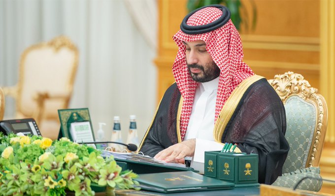 Saudi Crown Prince Mohammed bin Salman, wearing traditional red-checkered keffiyeh and bisht, reviews documents while chairing a Cabinet session in Riyadh, with official green-bound folders and ceremonial items arranged on the table before him.
