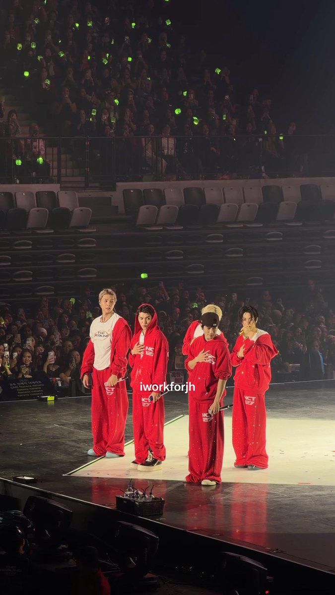 Four performers in matching red hooded tracksuits stand on stage with hands over their hearts, facing an audience holding green light sticks, with a watermark reading 'iworkforjh'.