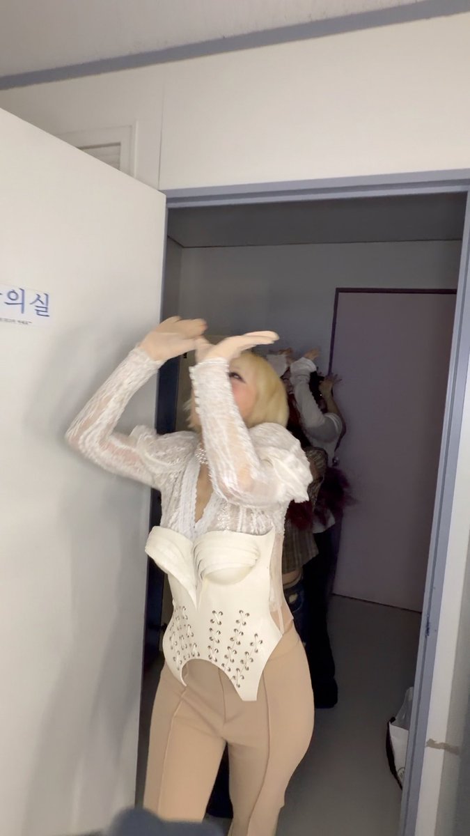 A person in a blonde wig, white lace top, white corset with rhinestone details, and beige leggings poses in a backstage hallway with Korean text on the door.