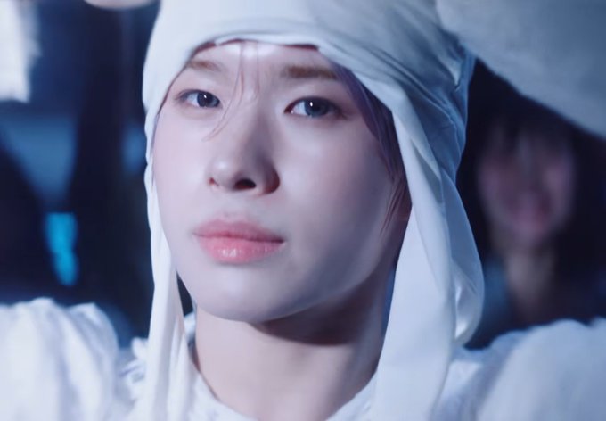 Close-up of a young woman wearing a white head covering, looking thoughtfully off-camera in a softly lit setting.