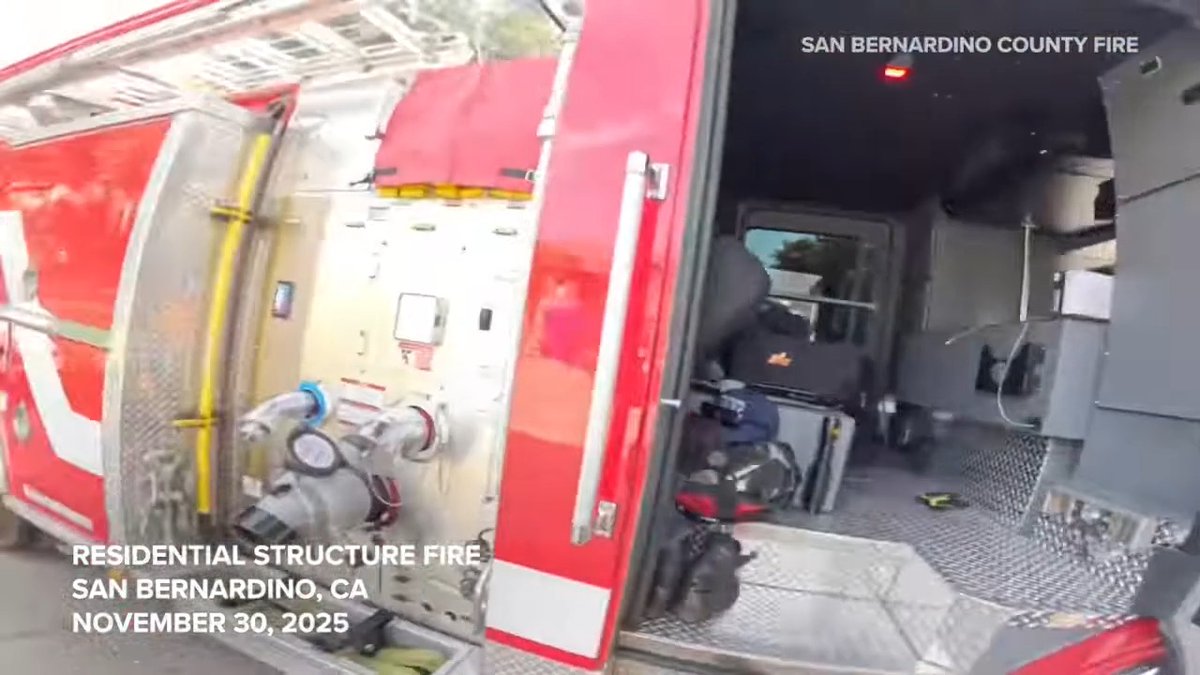 A frame from body camera footage showing the open compartment of a fire truck with text overlays: 'SAN BERNARDINO COUNTY FIRE' and 'RESIDENTIAL STRUCTURE FIRE SAN BERNARDINO, CA NOVEMBER 30, 2025'.