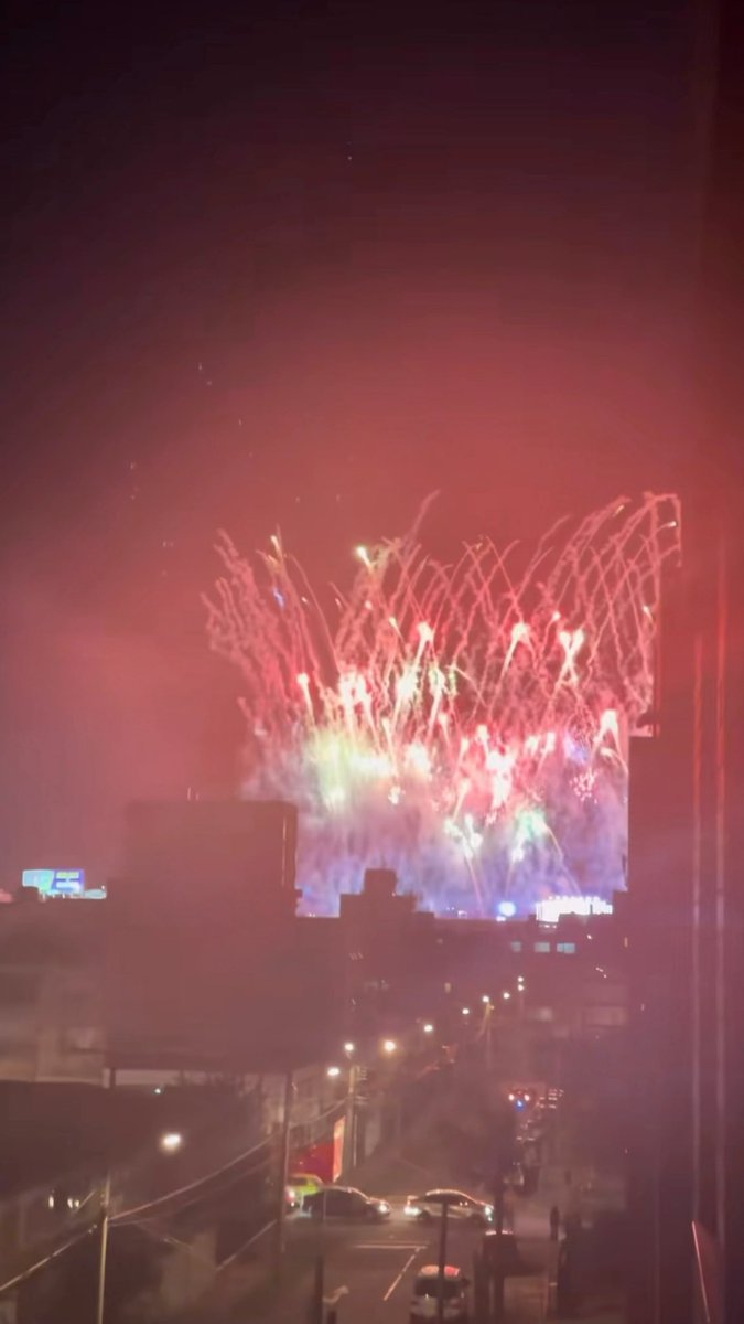 A nighttime photograph showing a large building illuminated by an impressive fireworks display in red, pink, and blue colors, with a street and cars visible in the lower foreground.