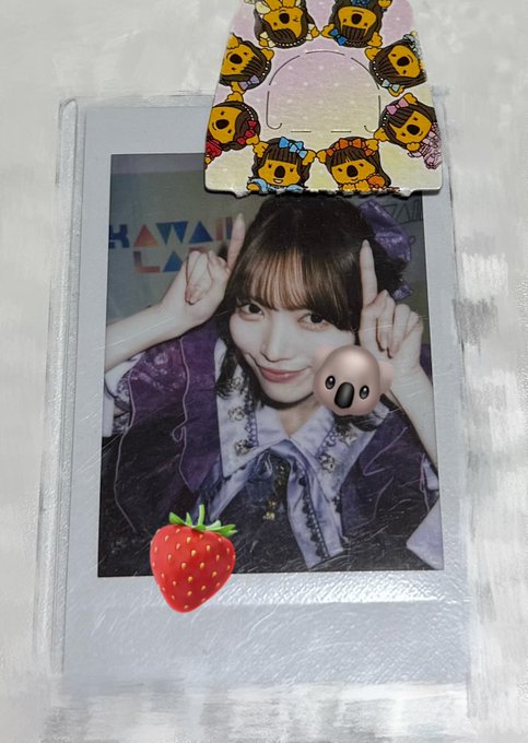 A photocard featuring a young woman posing with her fingers raised near her face, decorated with cartoon stickers and a strawberry emoji, displayed on a surface with a character-themed clip above it.