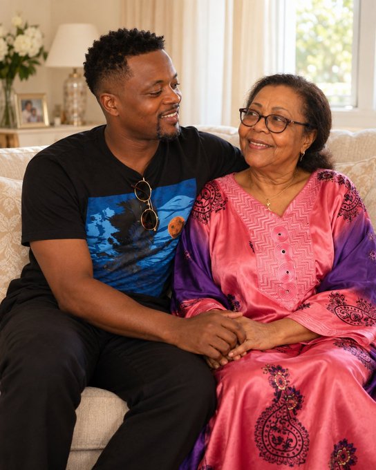 A man wearing a graphic t-shirt and sunglasses on his collar sits next to an older woman in a pink dress on a couch.