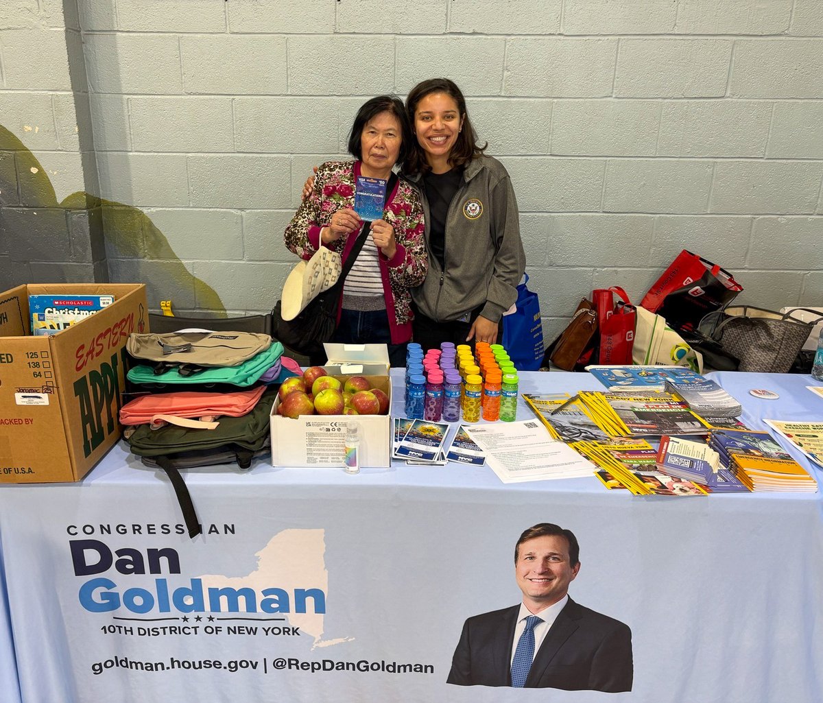 Two women pose behind a community information table, with one holding up a card. The table displays fruit, reusable bags, water bottles, and stacks of informational materials. A campaign banner with a man's portrait hangs across the front. There is text throughout the image.