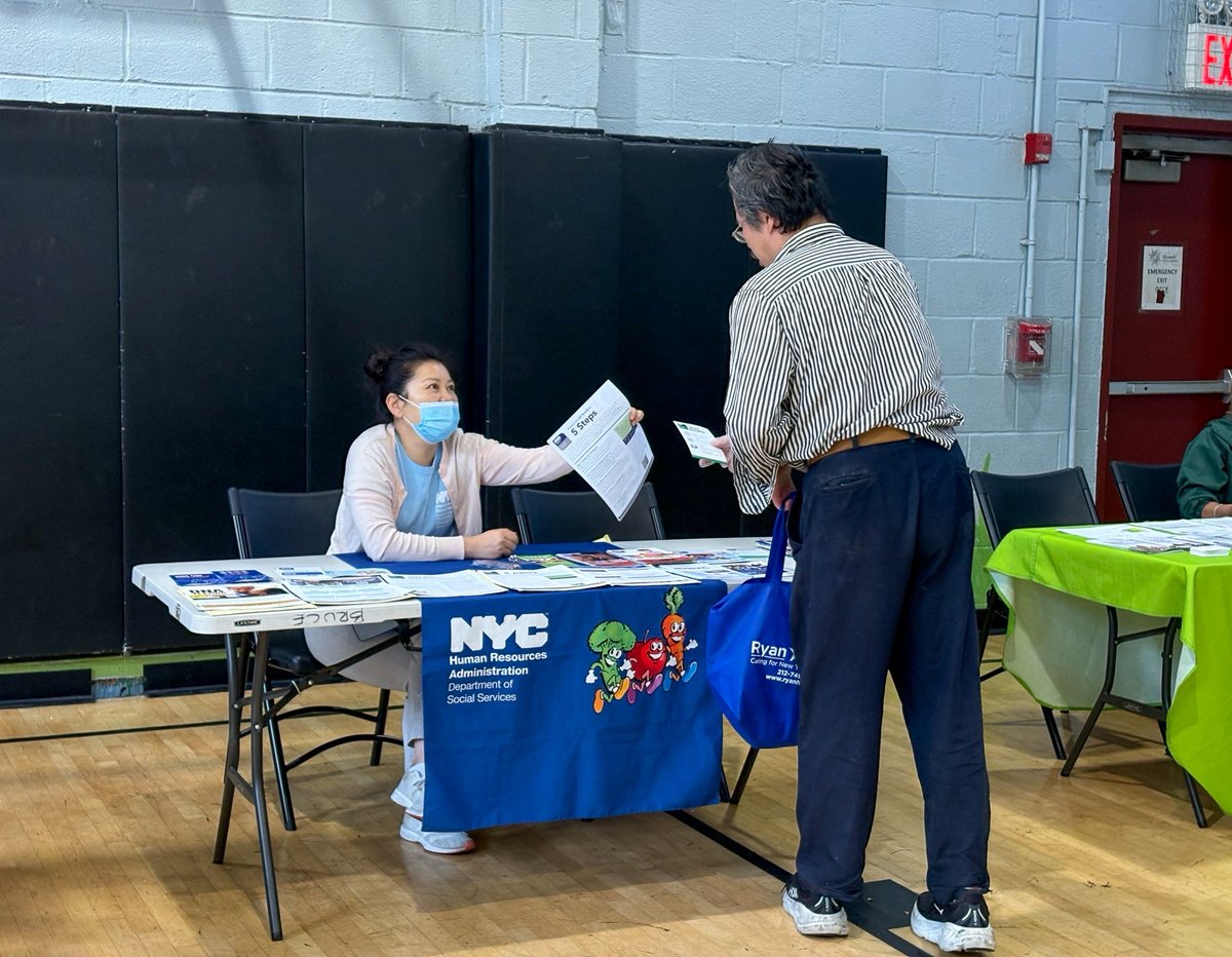 A masked woman seated at an information table hands a brochure to a man standing in front of her at an indoor community event. Brochures and papers cover the table, which has a blue department banner; another green-covered table and a doorway are visible nearby. There is text on the banner, brochures, and signs.