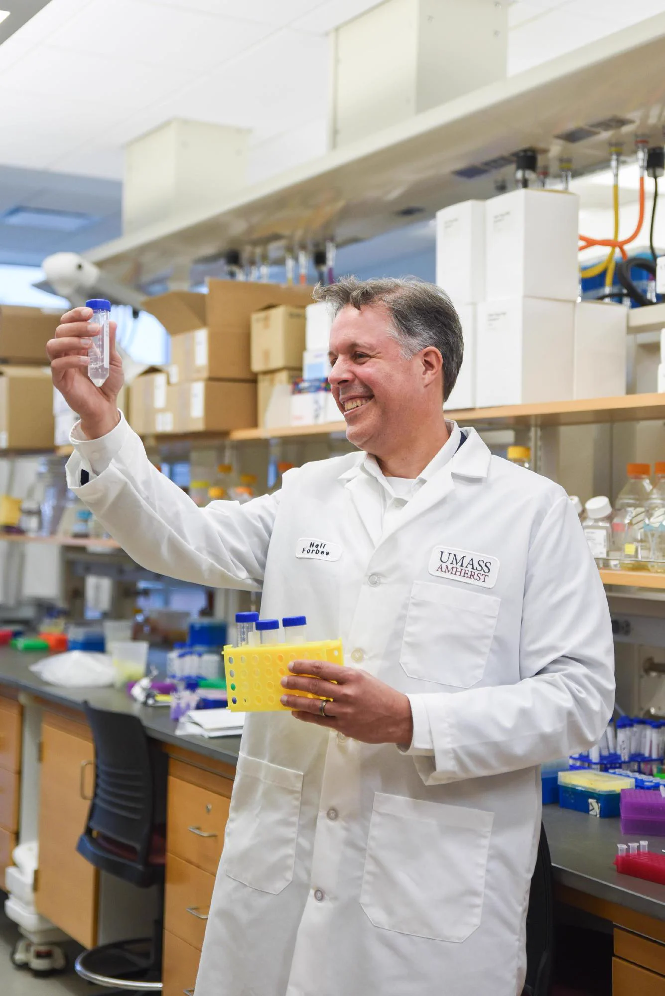 A person stands in a laboratory setting, wearing a white lab coat. They are holding a yellow container of test tubes in one hand, and a singular test tube, holding it up to the light to look at it, in the other hand. There are shelves of laboratory equipment behind the person, and their lab coat says "Neil Forbes" and "UMass Amherst" in small text on either side above the chest pockets.