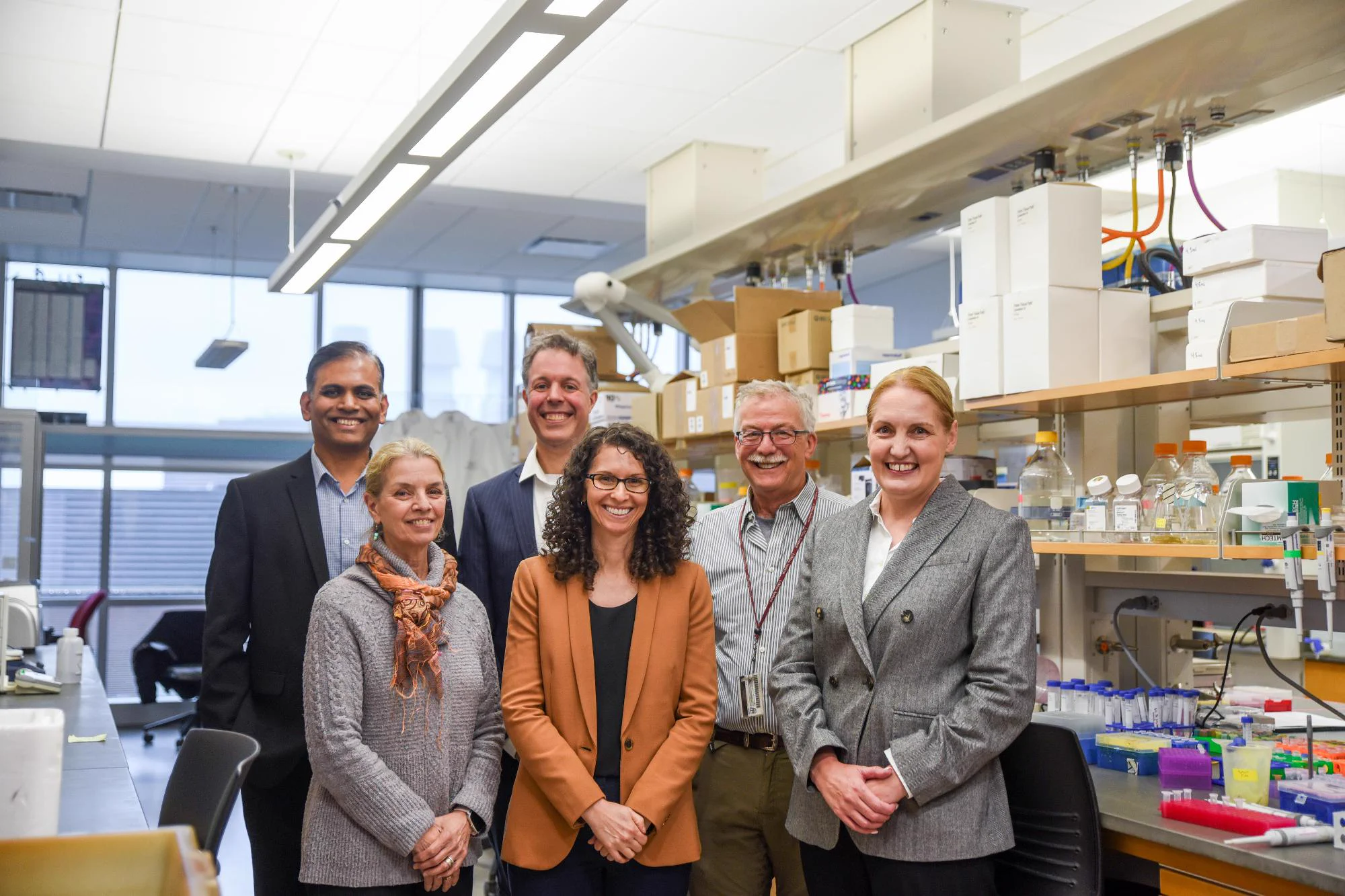A group of six people stand and smile at the camera in a laboratory setting. There are windows and shelves of laboratory equipment behind the group, and bright overhead lights.
