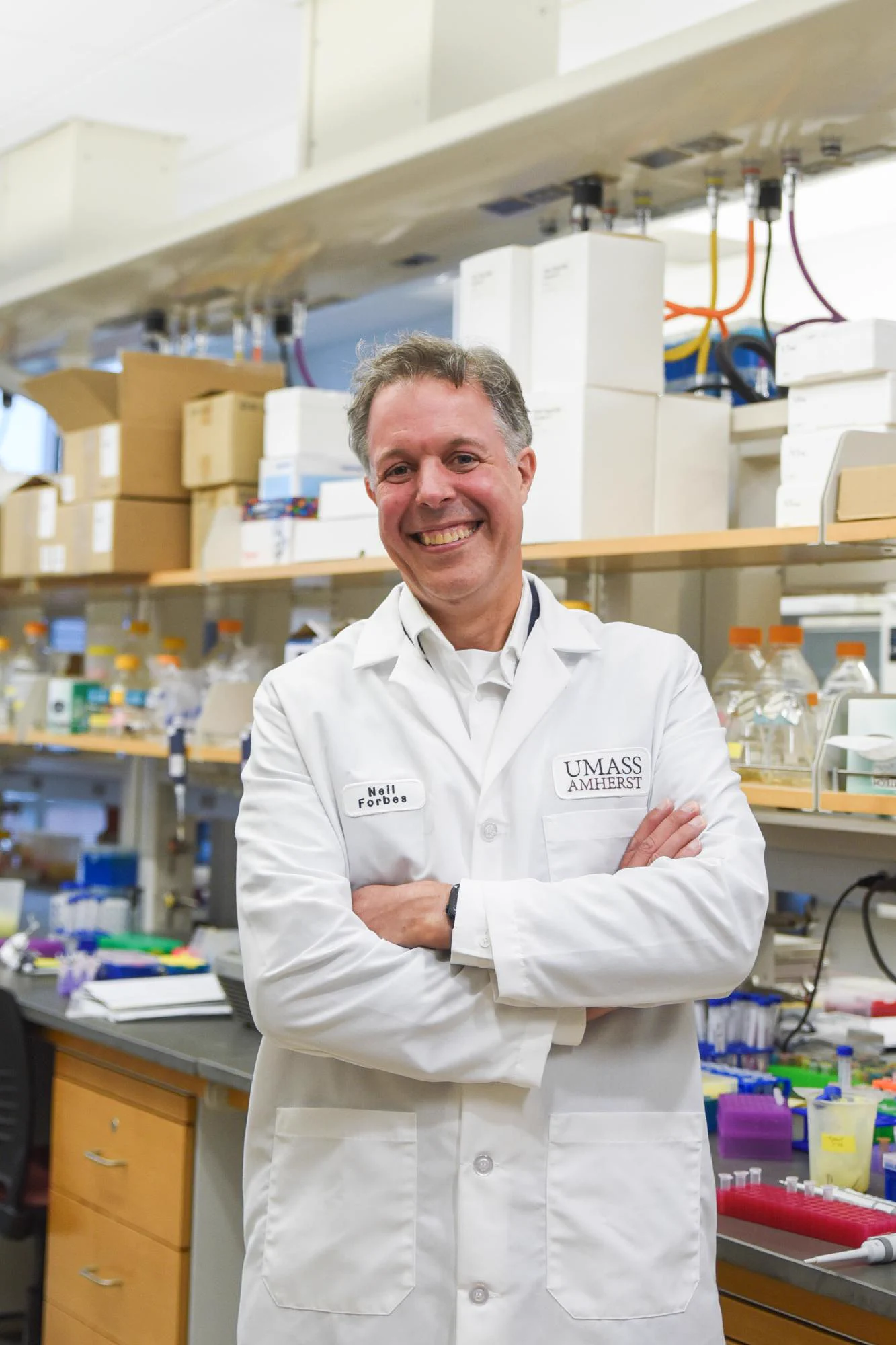 A person stands in a laboratory setting, wearing a white lab coat, crossing their arms and smiling at the camera. There are shelves of laboratory equipment behind the person, and their lab coat says "Neil Forbes" and "UMass Amherst" in small text on either side above the chest pockets.