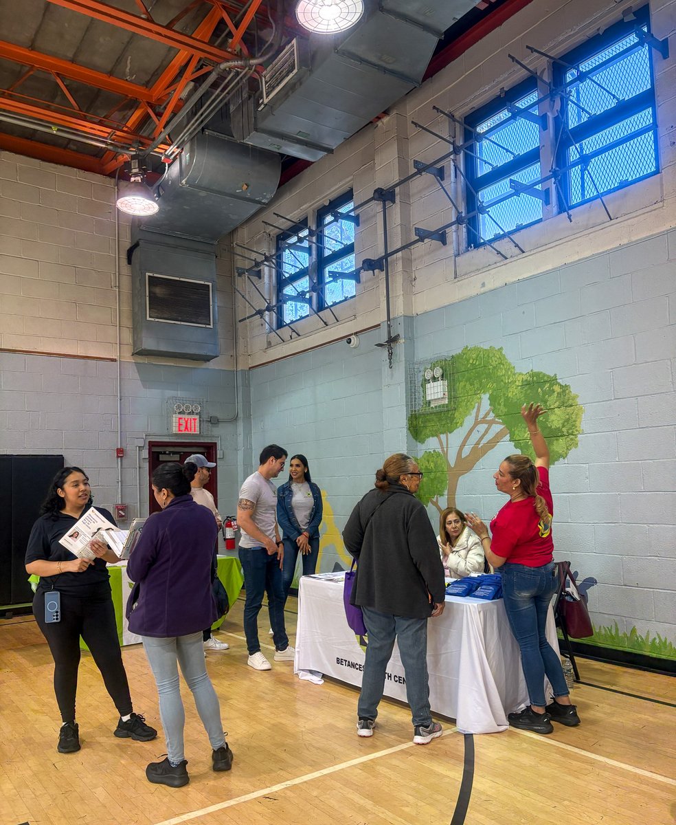 People gather around information tables at an event in a gymnasium. One woman holds a brochure while another speaks to a group, and a person behind a table gestures with a raised hand. The room has high windows, exposed ceiling fixtures, and a painted tree mural. Some text is visible on a brochure and table covering.