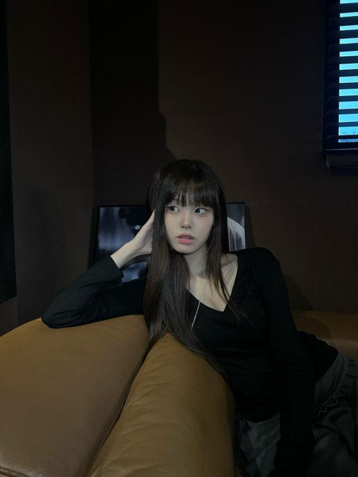 A young woman with long dark hair and bangs, wearing a black top, poses with her hand near her face while sitting on a tan leather couch.