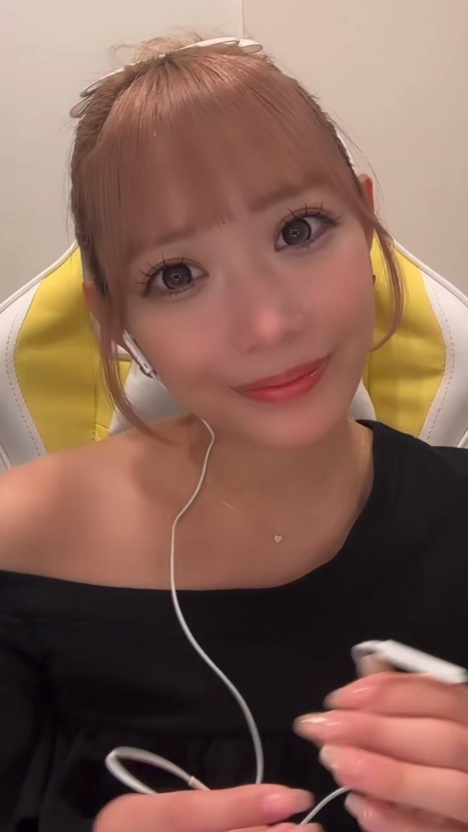A selfie-style portrait of a young woman with light brown hair and bangs, wearing a black top, red lipstick, and white wired earphones, sitting in a yellow and white gaming chair.