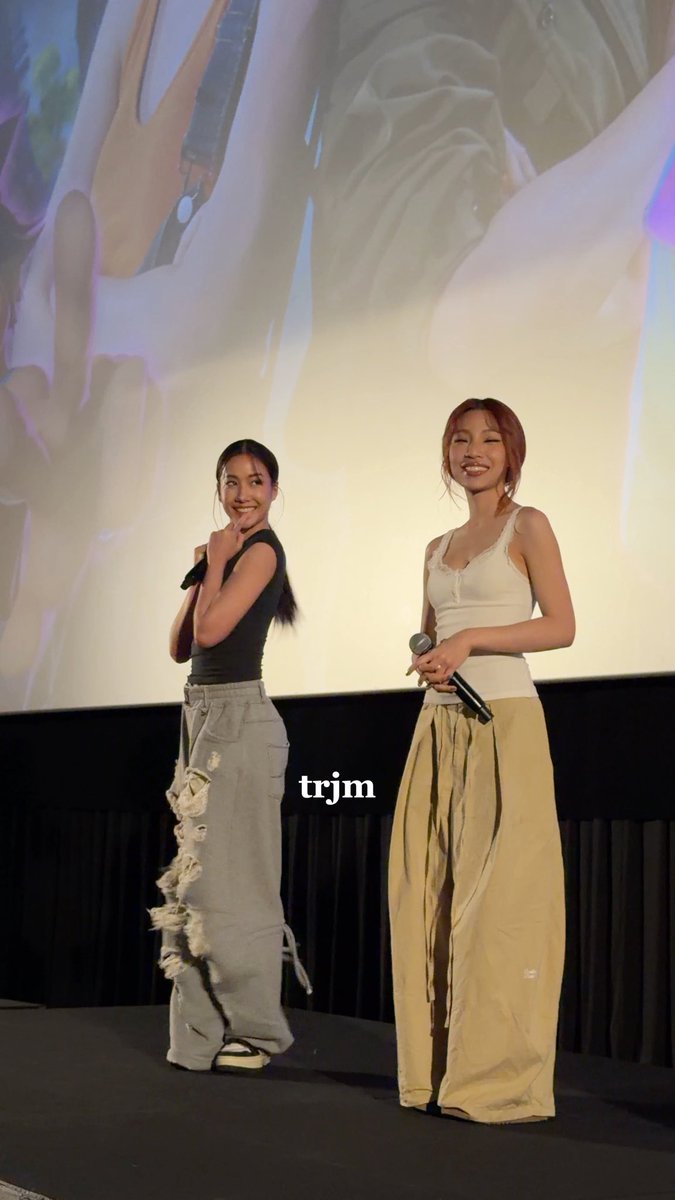 Two young women stand on a stage holding microphones; one wears a black tank top with distressed gray jeans, and the other wears a white tank top with tan cargo pants, with the text 'trjm' overlaid.