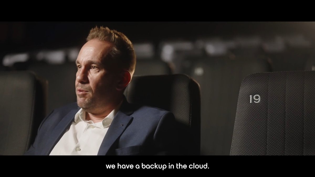A man in a white shirt and dark blazer sits in a dark theater seat marked "19" with the subtitle "we have a backup in the cloud."