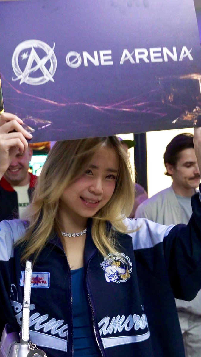 A young woman with blonde hair at what appears to be a gaming event, wearing a blue and white varsity jacket with patches and holding up a sign that reads 'ONE ARENA' with a circular logo.