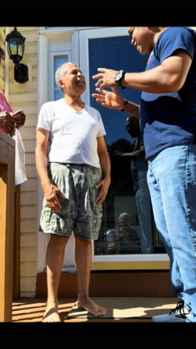 An outdoor photo showing an older man in a white t-shirt and shorts standing on a porch, listening to another person in a blue shirt who is gesturing with their hands.