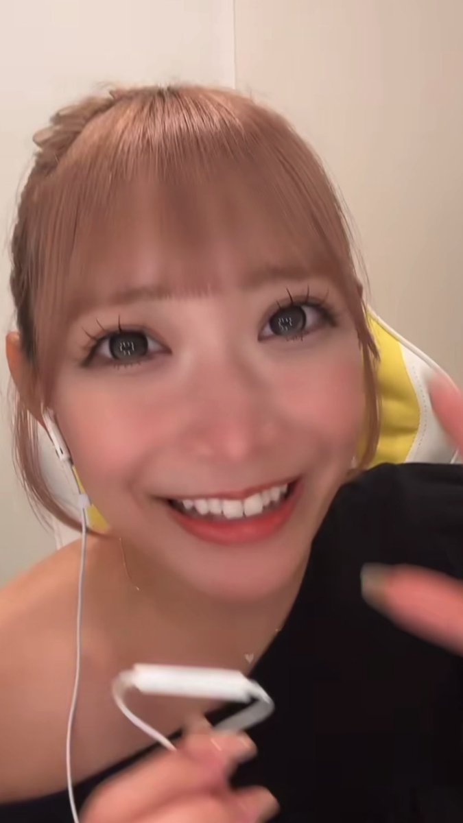 A close-up selfie of a smiling young woman with light brown hair, large eyes, and wearing a yellow and white headband, holding what appears to be a controller or cable.
