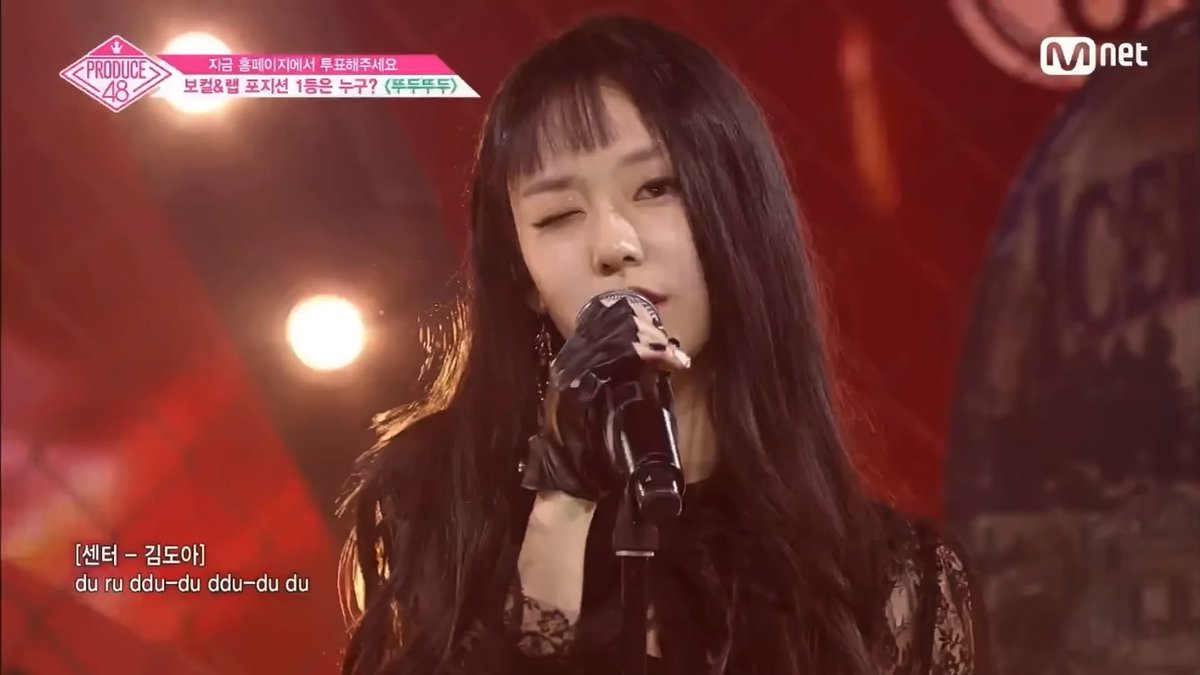 A performer sings into a microphone on a stage with a red background. The screen shows 'PRODUCE 48' logo, 'Mnet' channel logo, and text including '[센터 – 김도아]' and 'du ru ddu-du ddu-du du'.
