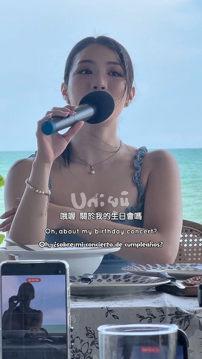 A young woman sits at a seaside dining table holding a small black fan to her mouth; the video includes subtitles in Thai, Traditional Chinese ('哦喔 關於我的生日會嗎'), and Spanish ('Oh, ¿sobre mi concierto de cumpleaños?').