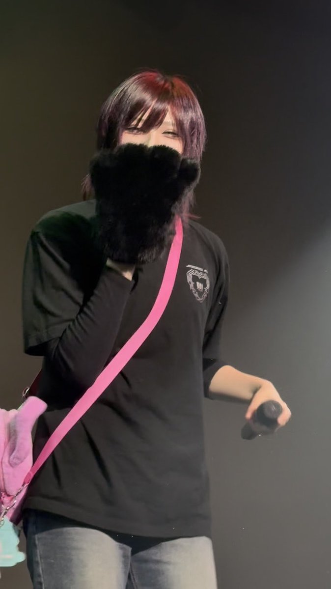 A person with reddish hair wearing a black shirt, large black furry gloves, and a pink crossbody bag holds a microphone on stage.