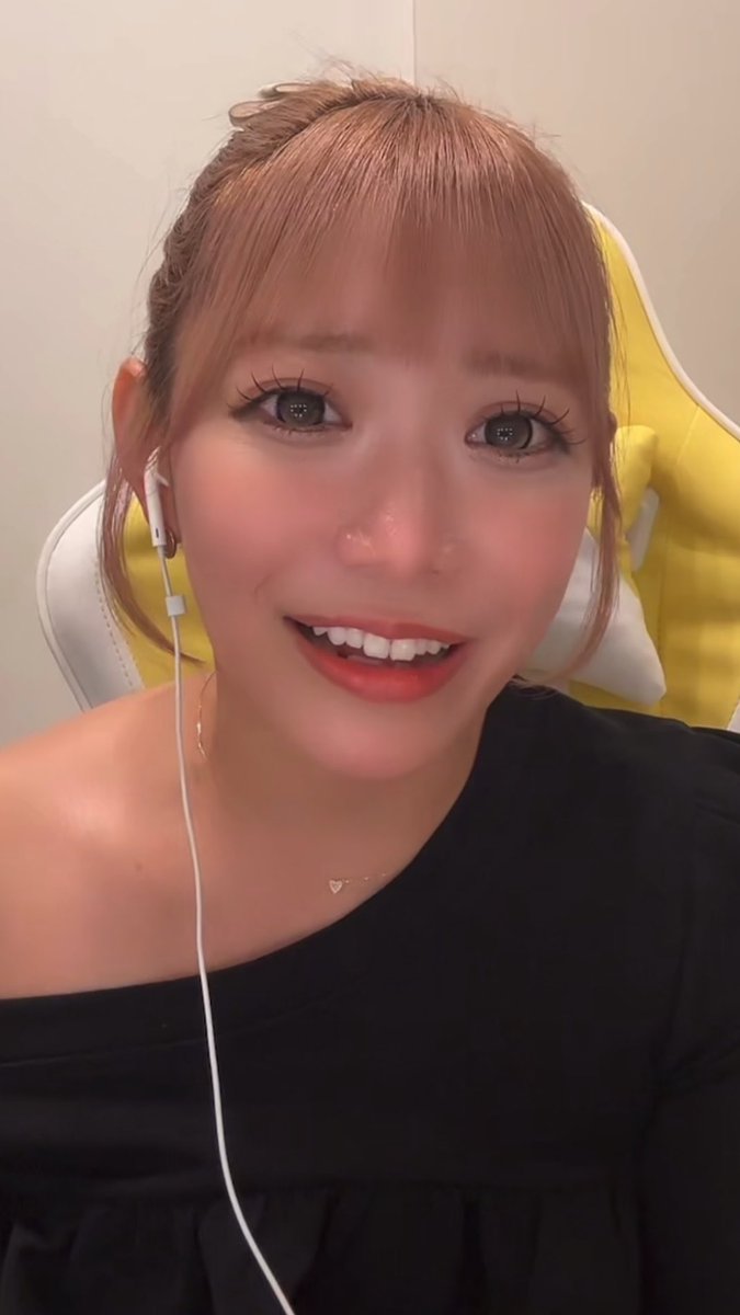A close-up selfie of a smiling woman with light brown hair and bangs, wearing red lipstick, a black off-shoulder top, and white wired earphones.