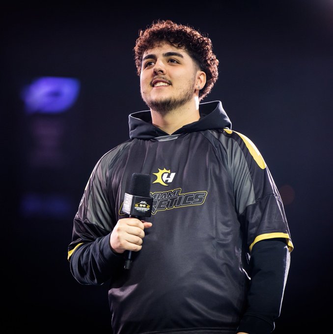 A curly-haired player wearing a Miami Heretics jersey holds a microphone with the Heretics logo, smiling slightly as he looks upward, fitting the announcement that ReeaL has officially rejoined Miami Heretics. The team's branding is clearly visible on his black and yellow hoodie-style jersey.