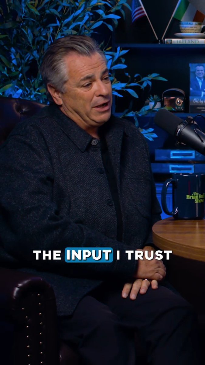 A man with graying hair wearing a black jacket seated in a studio setting, speaking into a microphone next to a 'The Brian Burns Show' mug, with the text overlay 'THE INPUT I TRUST'.